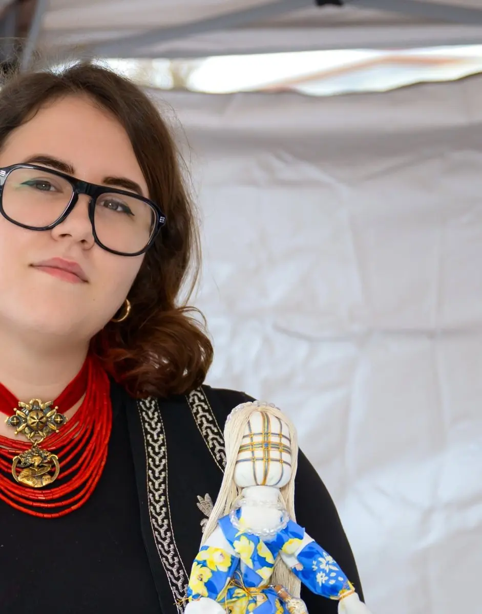 A community member wearing a red coral bead necklace holds a handmade Ukrainian motanka doll in a blue and yellow floral dress at UAFest