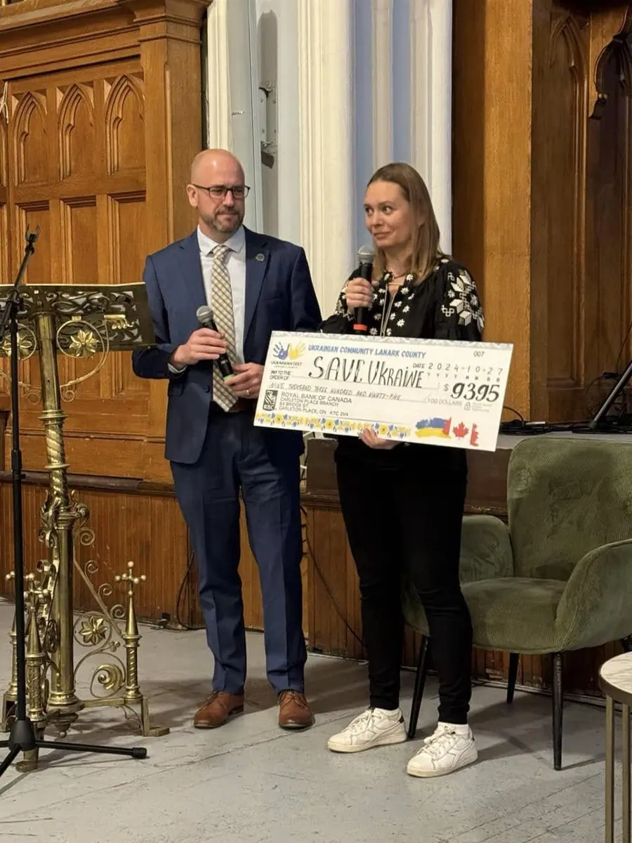A man in a suit and a woman in an embroidered blouse presenting a large ceremonial cheque for $9,395 to Save Ukraine at a formal indoor ceremony