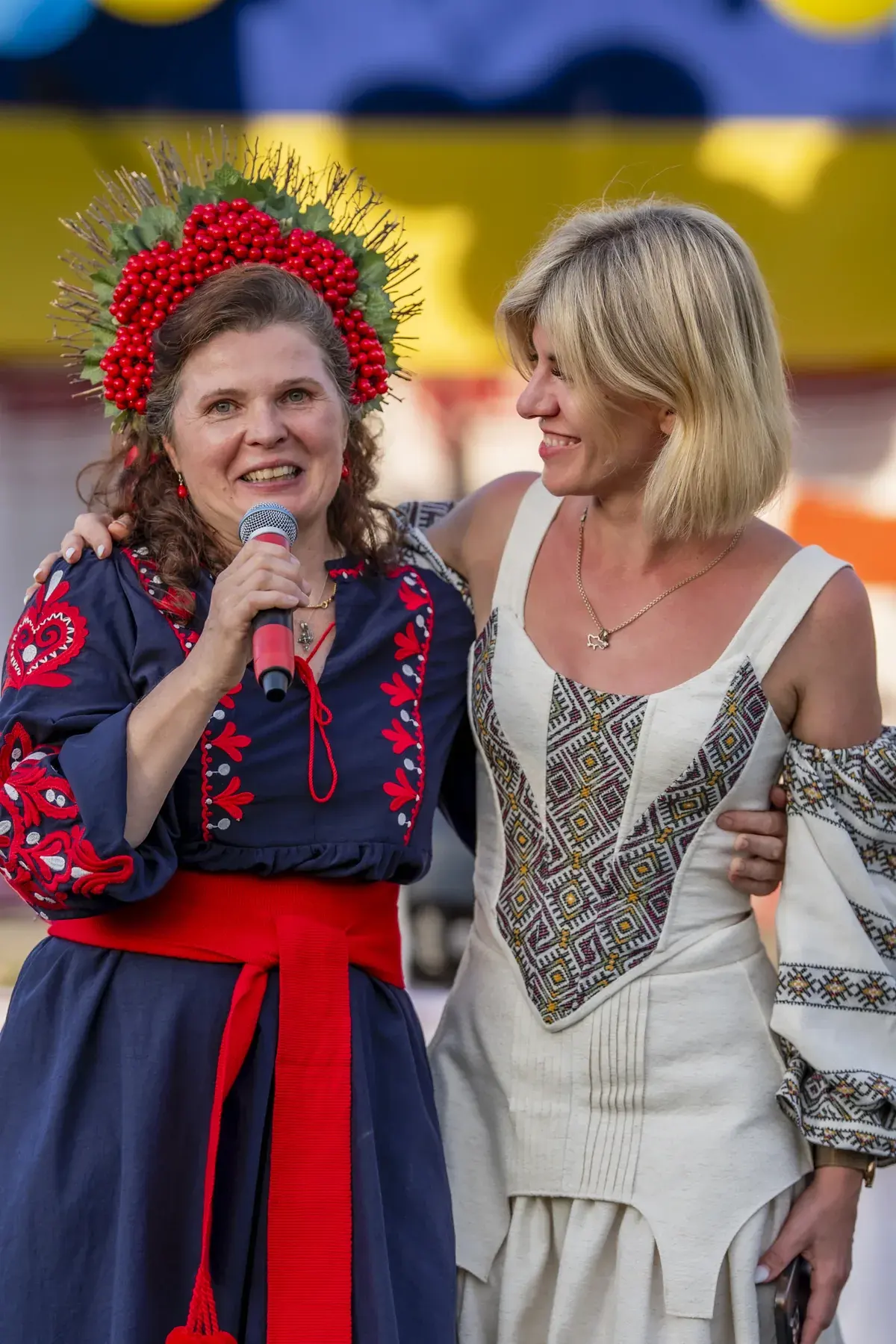 Oksana Pikush at UAFest 2026 — Woman in viburnum wreath speaks, embracing a blonde performer (photo: Tim Skinner)