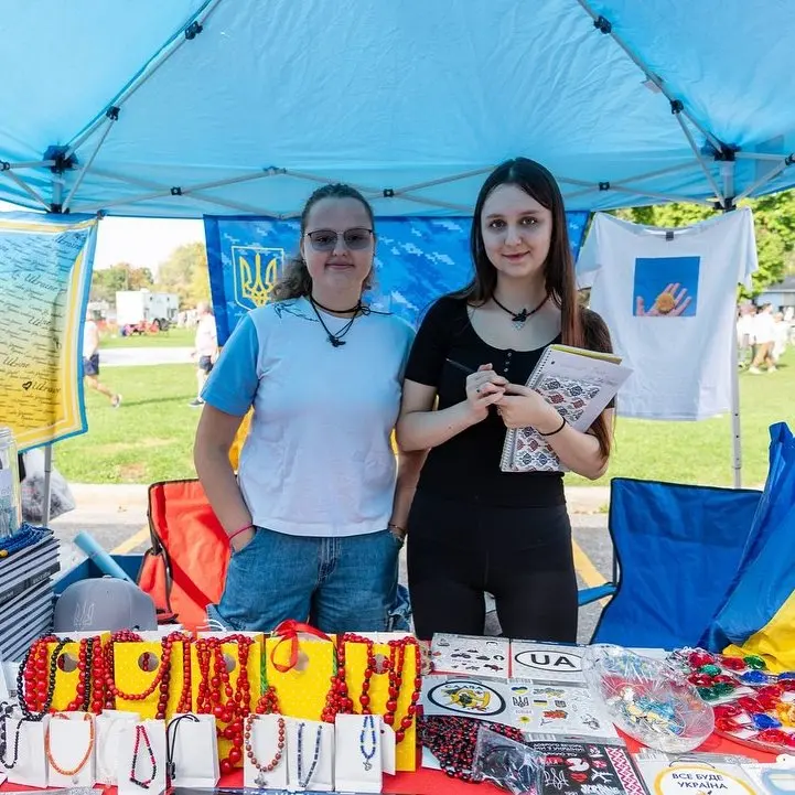 Two vendors behind their table under a blue tent at UAFest — jewellery, patches and embroidered pieces laid out on the stall, with the festival lawn behind them