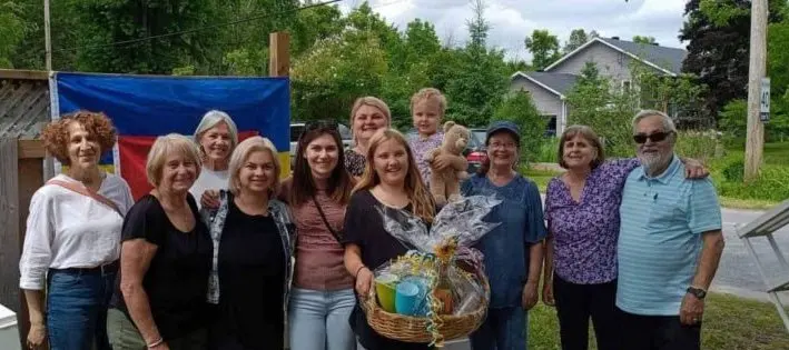 A group of UAFest organizers and supporters posing outdoors in front of a Ukrainian flag, holding a sunflower gift basket, at an early community fundraiser in Carleton Place