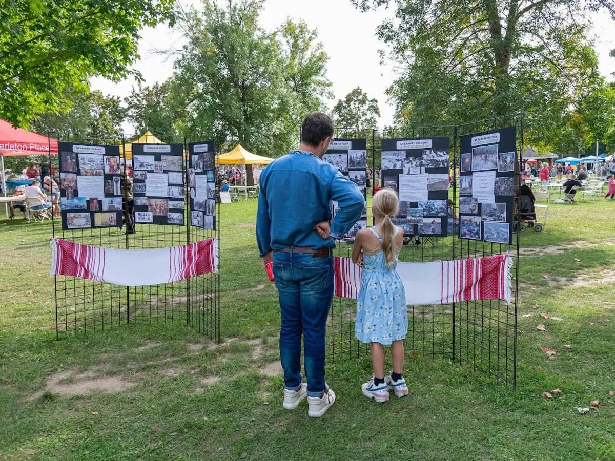 A visitor reading a "Ukrainian Ancestry" photo display board set up under the trees at the festival