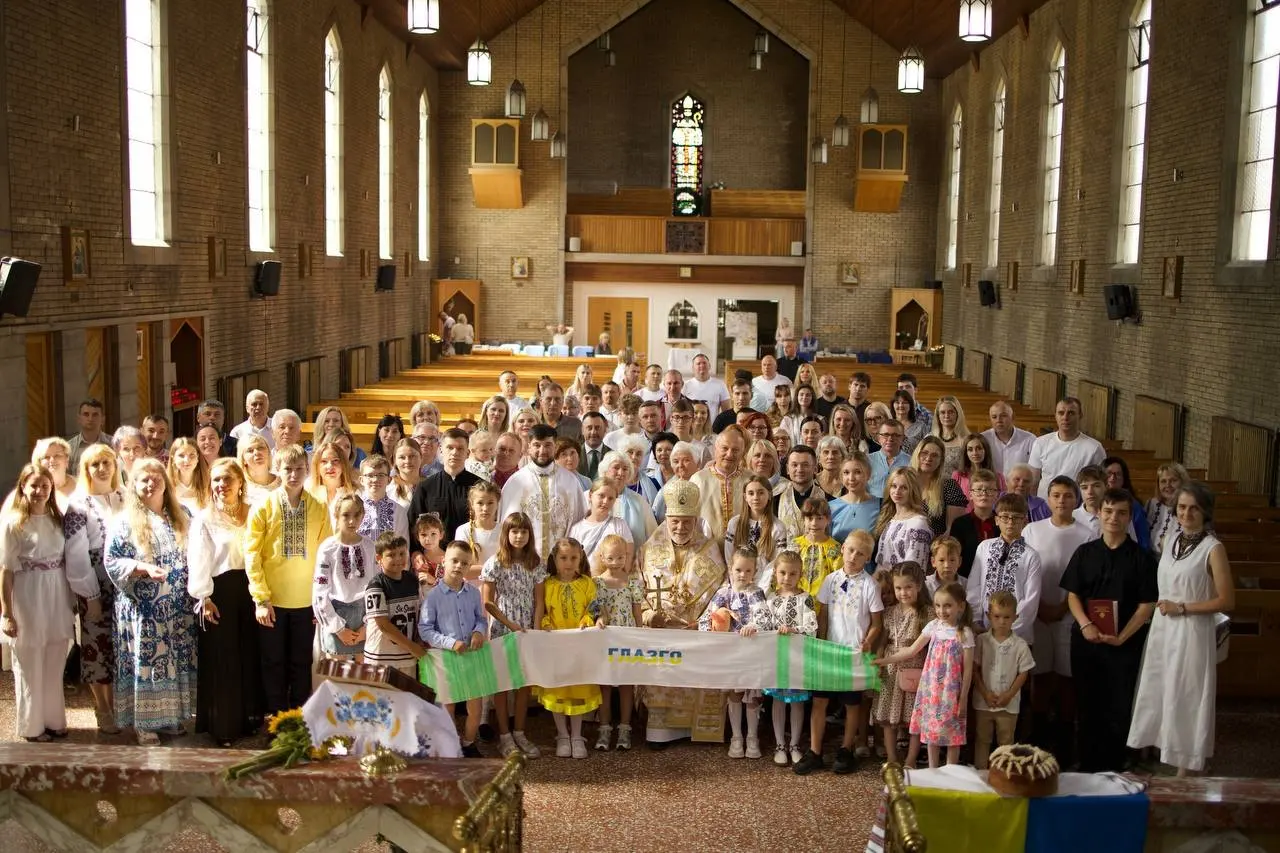 A large Ukrainian community gathered across the front of a church, many in embroidered vyshyvanky, holding a long embroidered panel marked ГЛАЗГО, with clergy at the centre