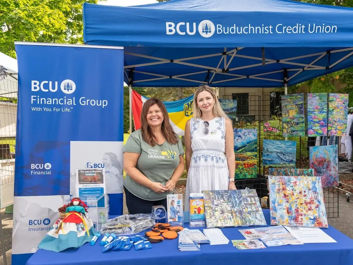 Two volunteers smiling at the Buduchnist Credit Union sponsor booth, with folk-art paintings displayed behind them.