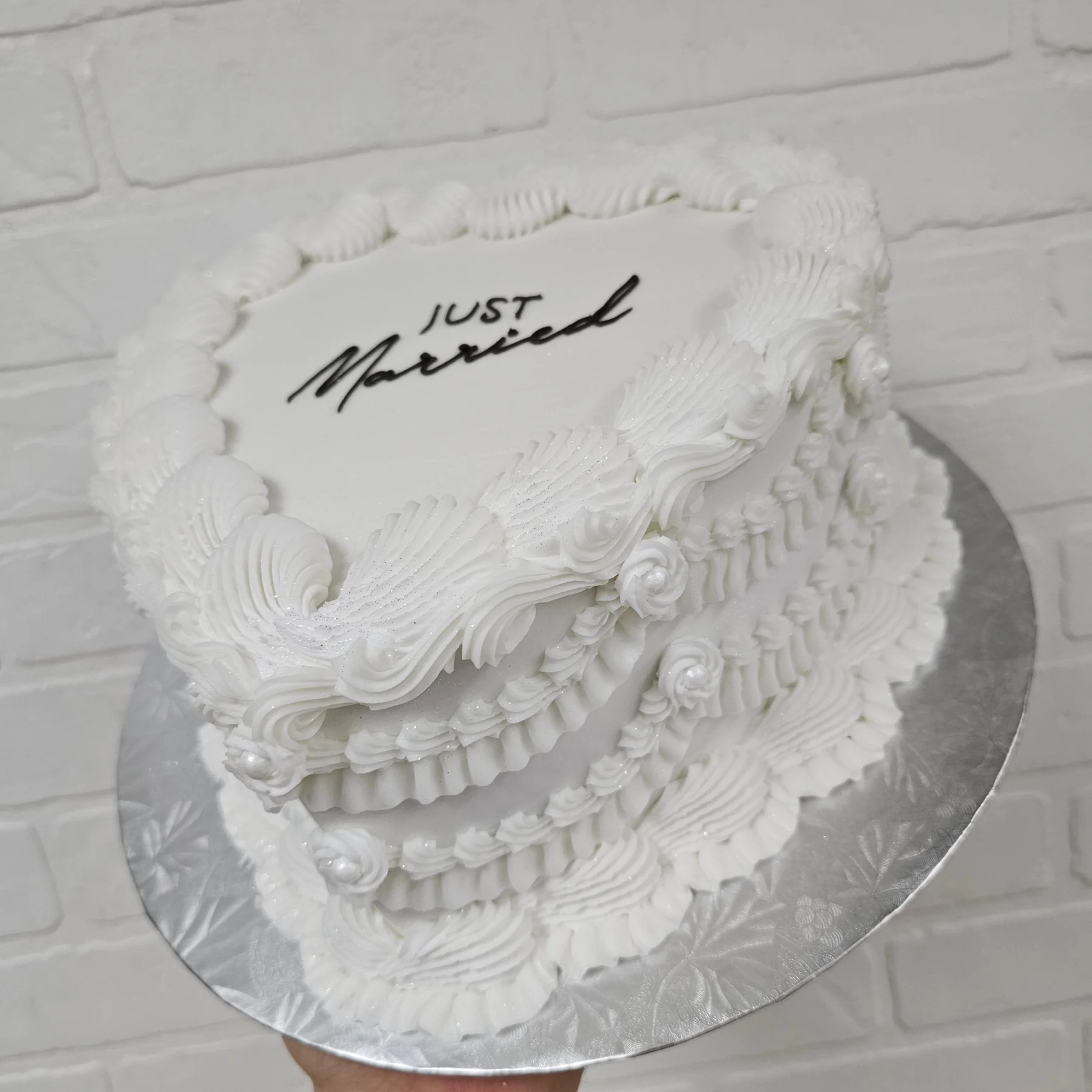 All-white heart-shaped Lambeth cake covered in piped shells, scrolls and pearls, with “Just Married” written on top