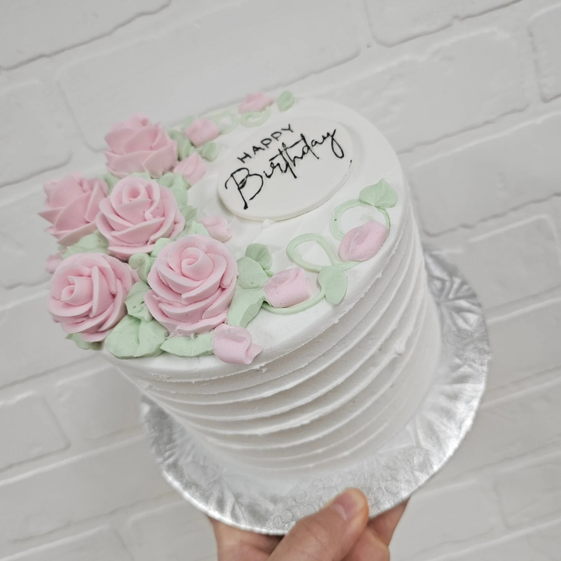Ribbed white buttercream cake topped with a cluster of piped pink roses, buds and green leaves beside a Happy Birthday plaque