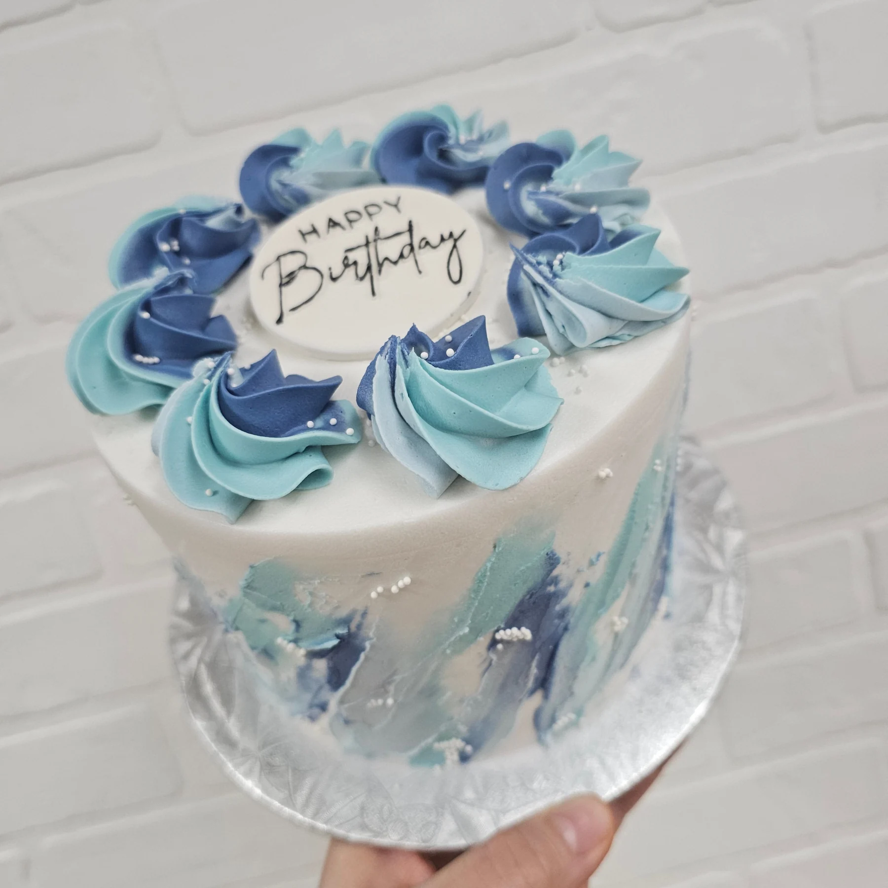 White cake with a blue palette-knife finish and a ring of blue and turquoise piped rosettes around a Happy Birthday plaque