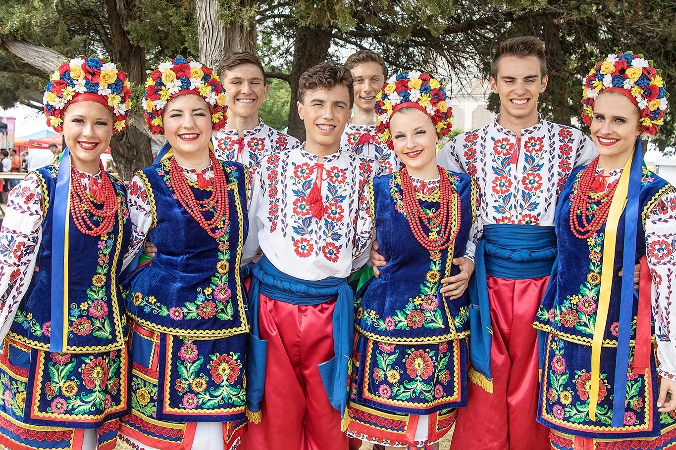 Young dancers in embroidered shirts, blue velvet vests, red sharovary and floral wreaths stand together outdoors at the festival