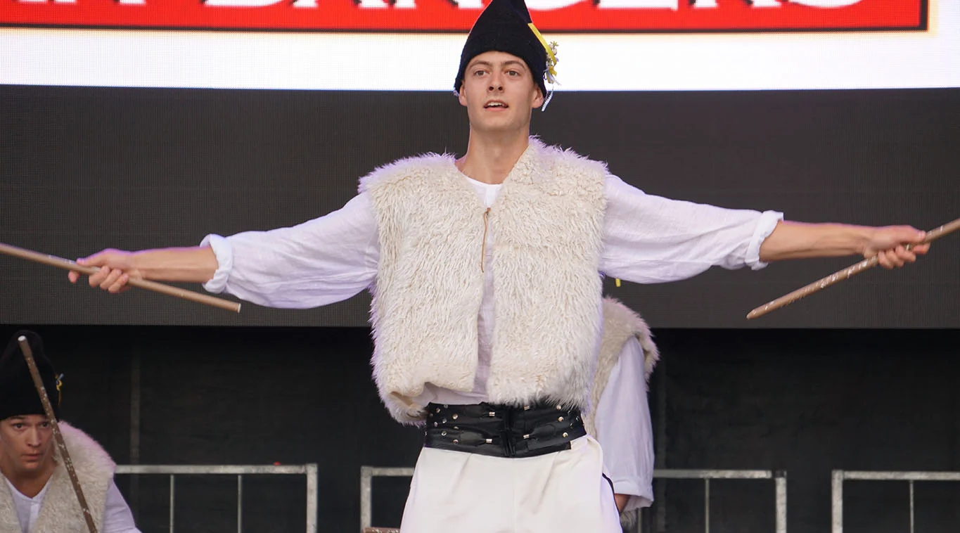 A dancer in a sheepskin vest and black felt hat holds two staffs wide apart on the festival stage