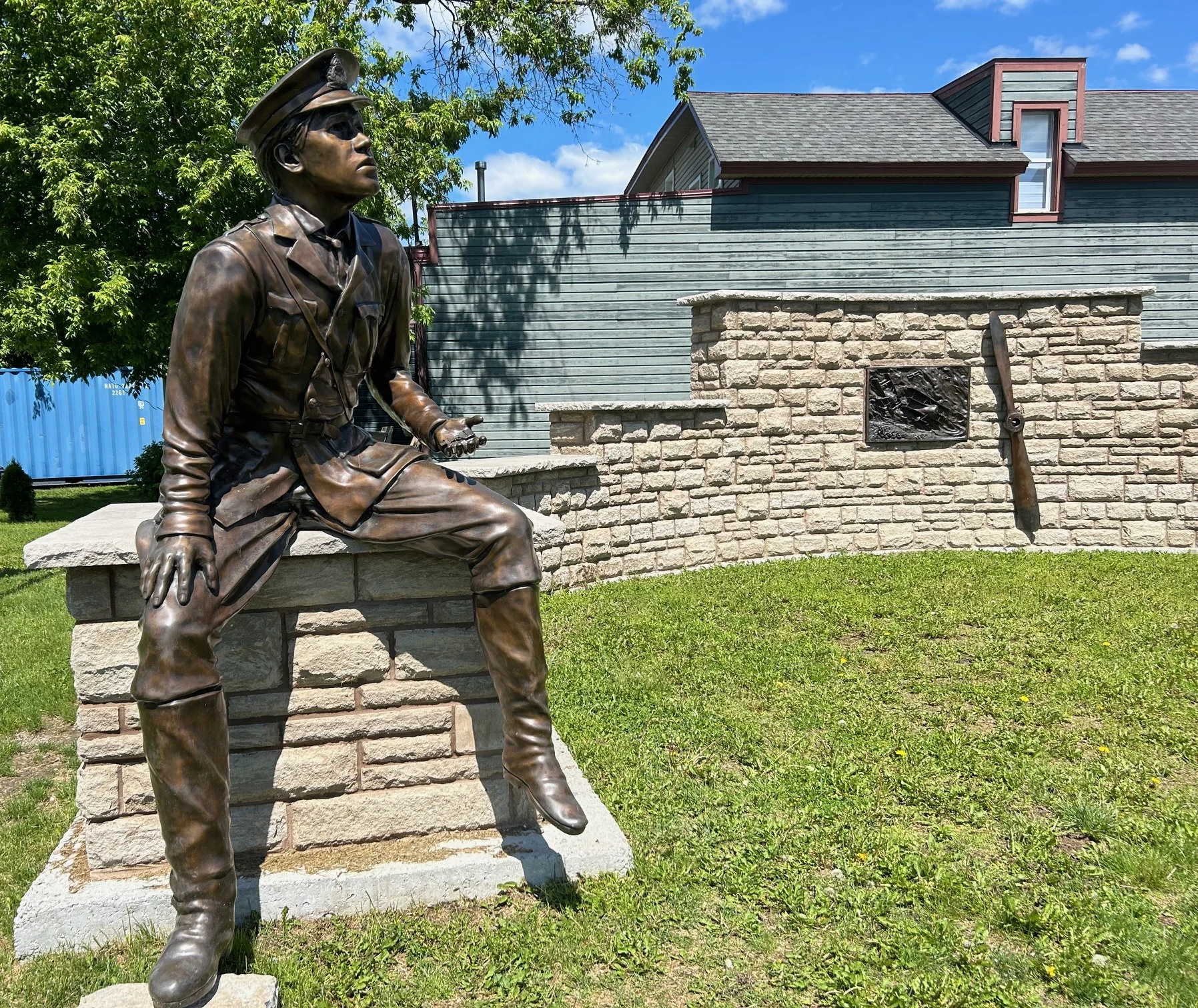 Bronze statue of Captain Roy Brown in flying uniform and peaked cap, seated on a stone plinth, with a propeller mounted on the stone wall behind him