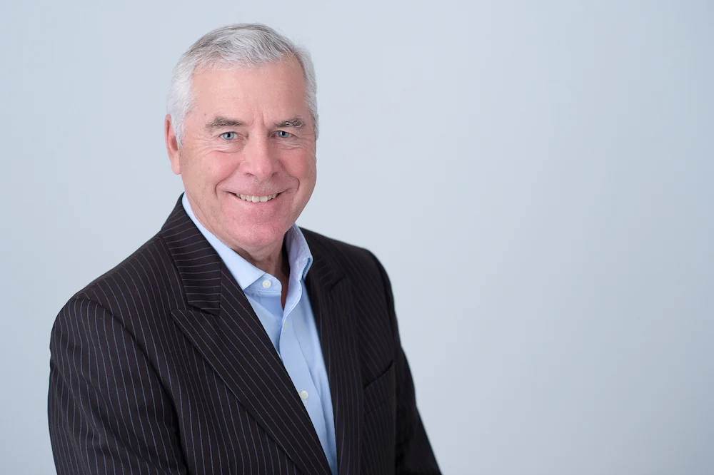 Marty Coulas, Vice President of Coulas Insurance, smiling in a dark pinstriped jacket and light blue open-collar shirt against a pale grey studio wall.
