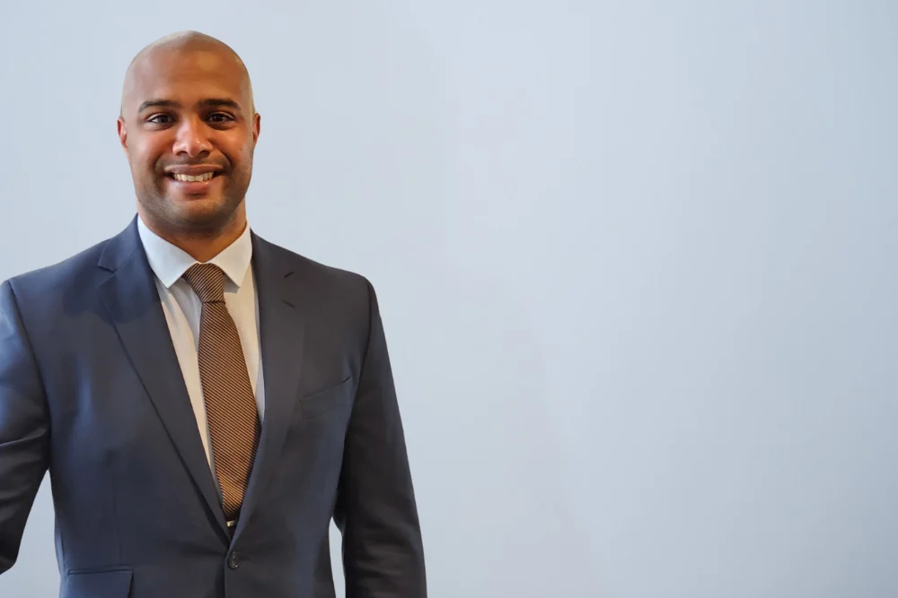 Samuel Ileka-Priouzeau, Director of Client Services at Coulas Insurance, smiling in a navy suit and brown striped tie against a pale grey studio wall.