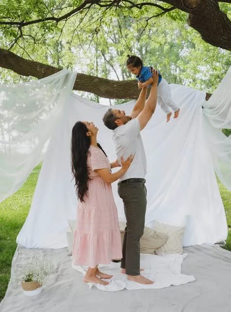 An outdoor family session of hers — a father lifting a toddler overhead beside the mother, in front of white drapes strung between trees
