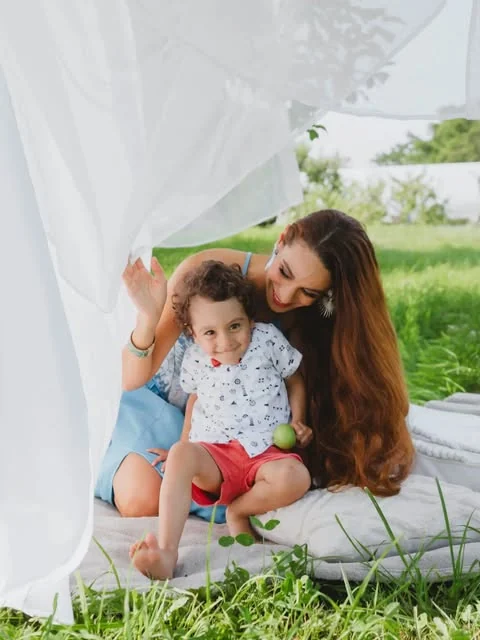 An outdoor session of hers — a mother lifting a white drape to look at her small son, sitting on a blanket in the grass