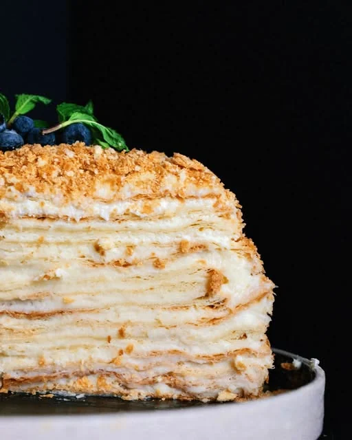 A Napoleon cake cut open to show its many pastry layers and cream, topped with crumbs, blueberries and mint