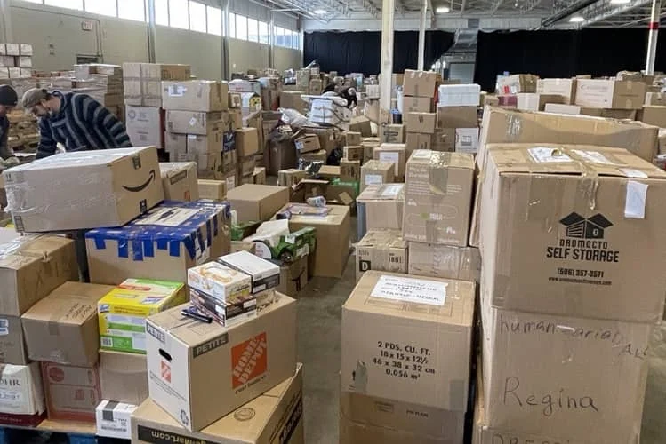 Volunteers sorting donated boxes on the warehouse floor, one crate hand-marked "humanitarian aid" and "Regina"