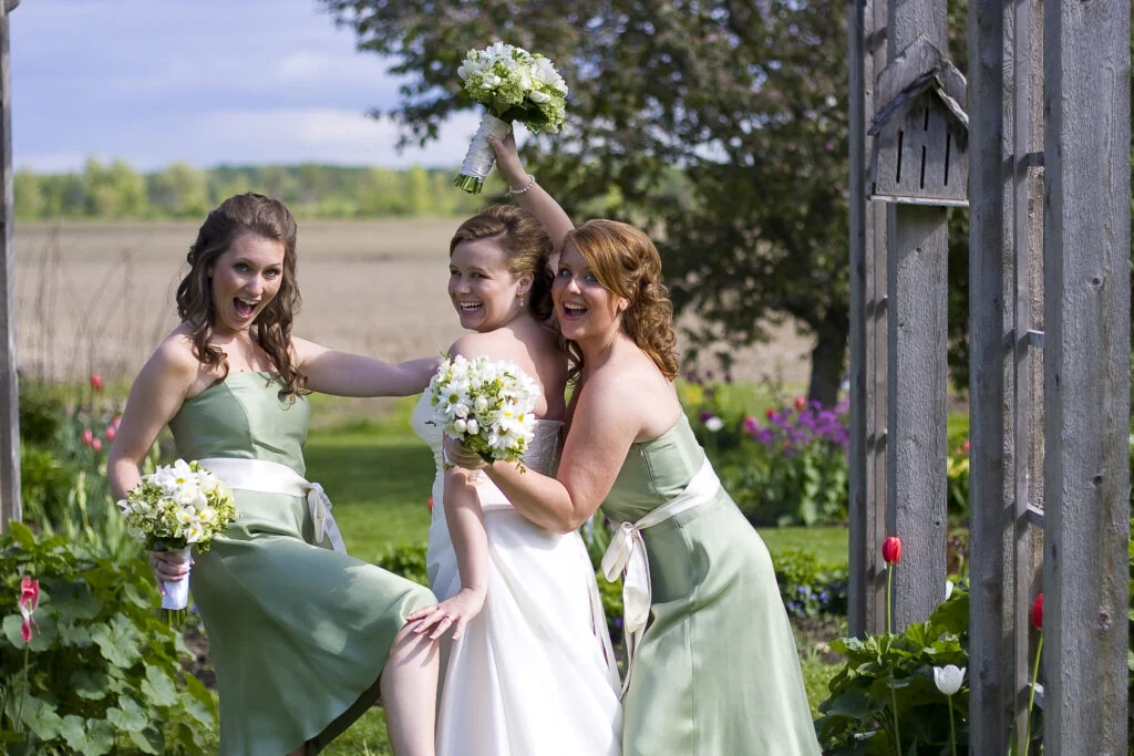 A laughing bride with a daisy bouquet held up by two bridesmaids in sage green dresses beside a wooden arbour and a spring garden