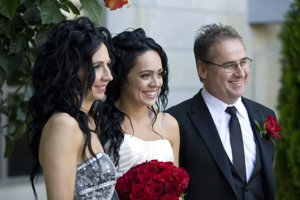 A bride with red roses stands between two family members outside a stone building, all three looking off camera and smiling