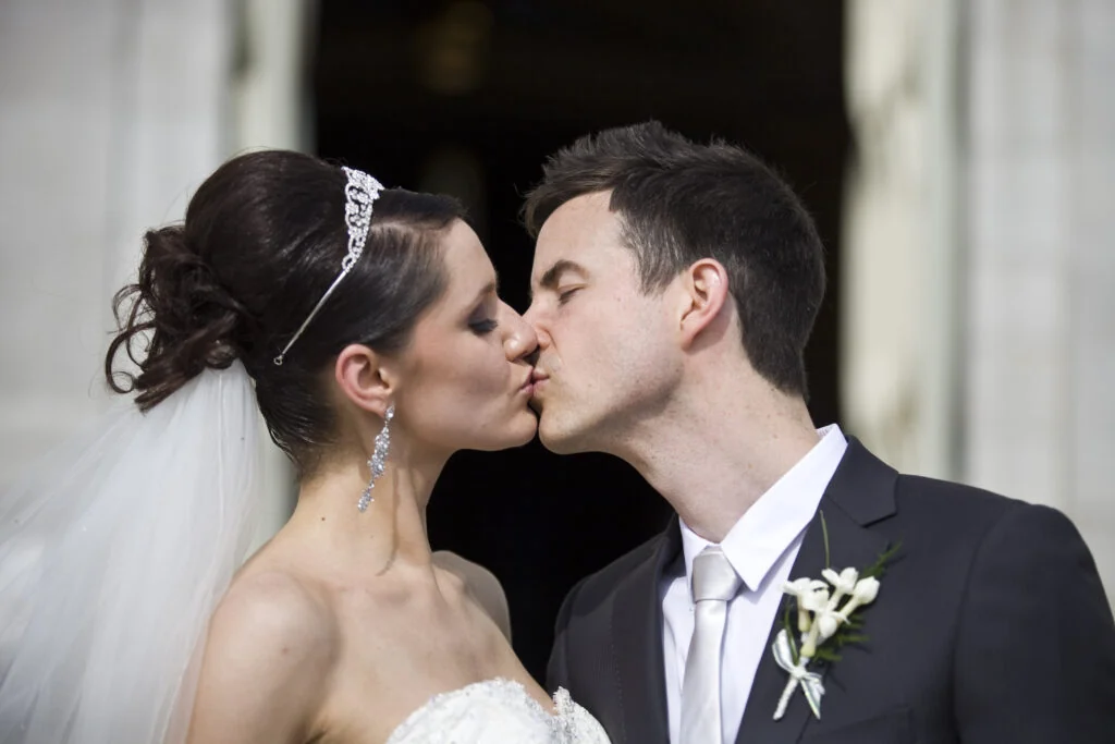 A bride in a tiara and veil and a groom in a dark suit kissing in front of a stone building