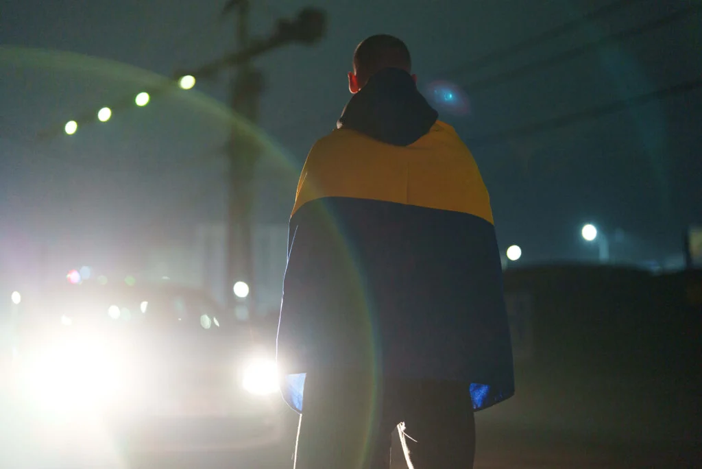 A young person seen from behind at night, wrapped in a Ukrainian flag, headlights ahead on the road