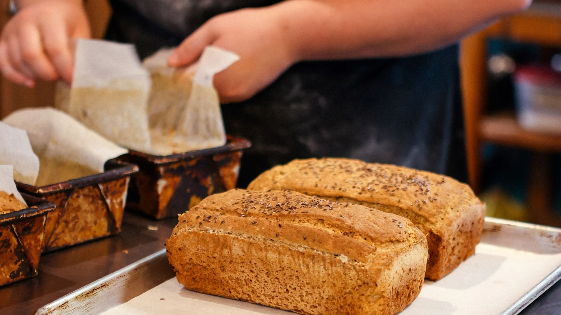 Two freshly baked loaves topped with flax seeds on a tray, with a baker lifting parchment from loaf tins behind them