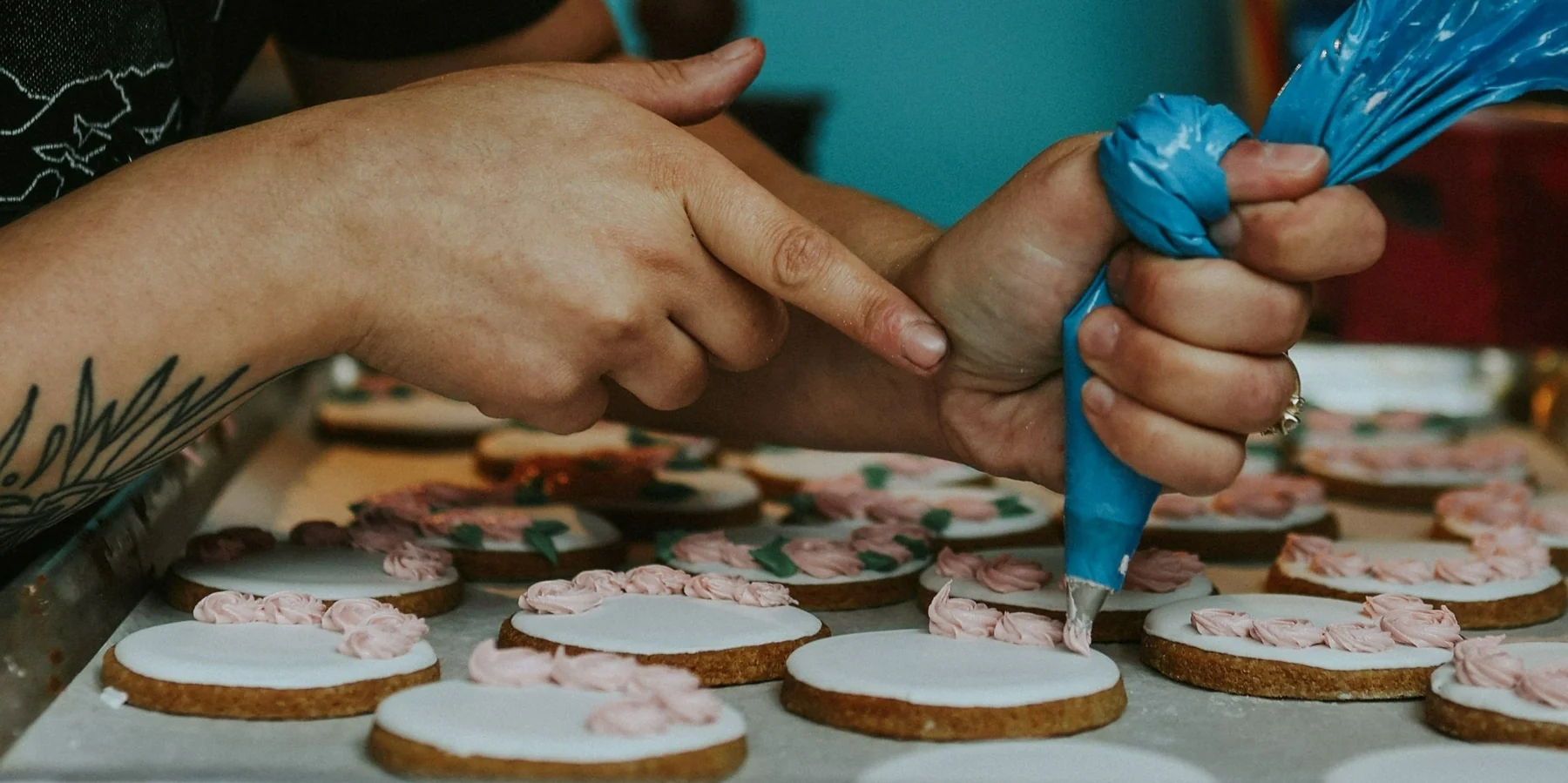 Two pairs of hands piping pink buttercream roses onto white-iced sugar cookies laid out on a baking sheet