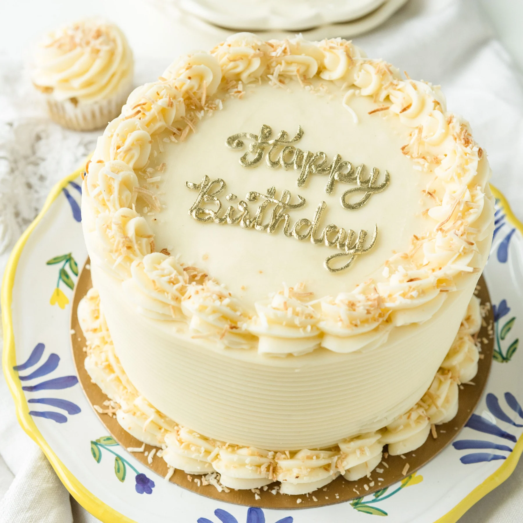 A round cake in ivory buttercream with a piped shell border, toasted coconut and a gold Happy Birthday topper, on a floral plate with a matching cupcake