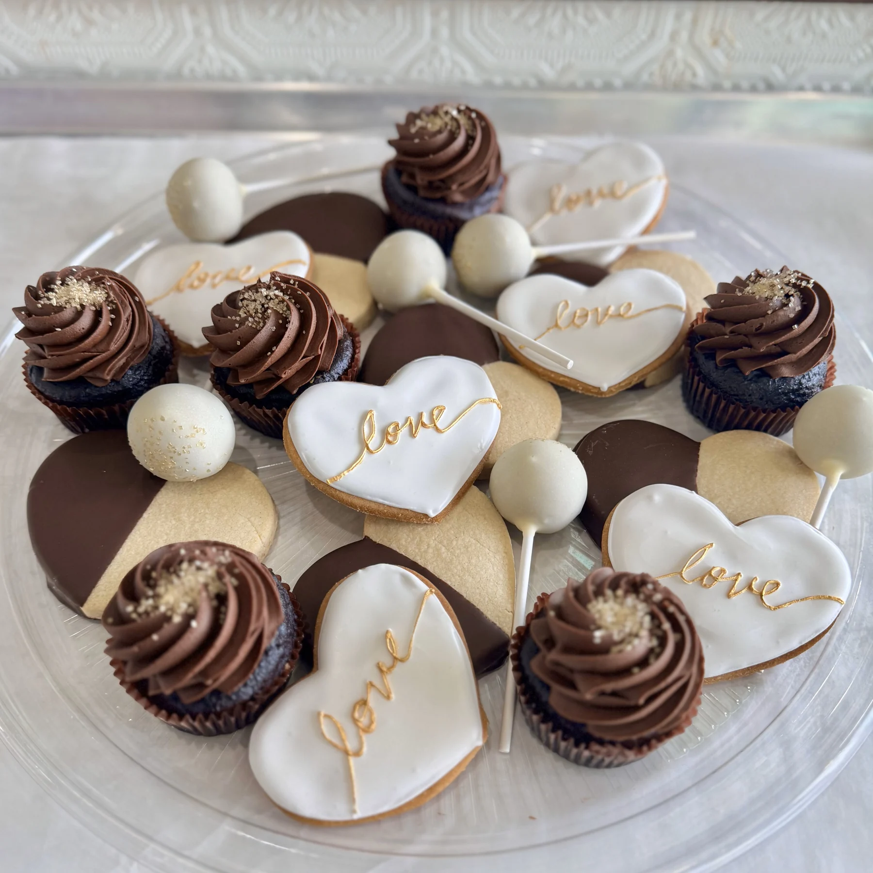 The wedding platter — heart-shaped sugar cookies iced in white with gold Love lettering, chocolate-dipped shortbread, chocolate cupcakes and white cake pops on a glass plate