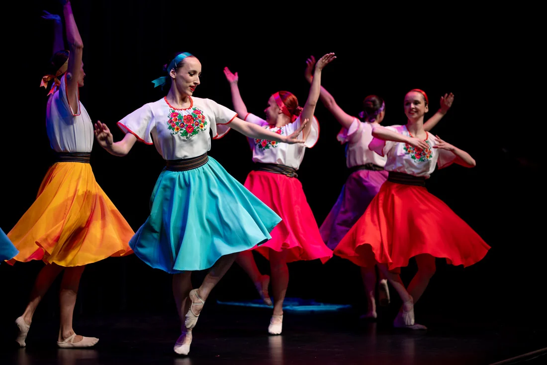 Five dancers in white embroidered blouses and full skirts of yellow, turquoise, pink and red turning on a darkened stage