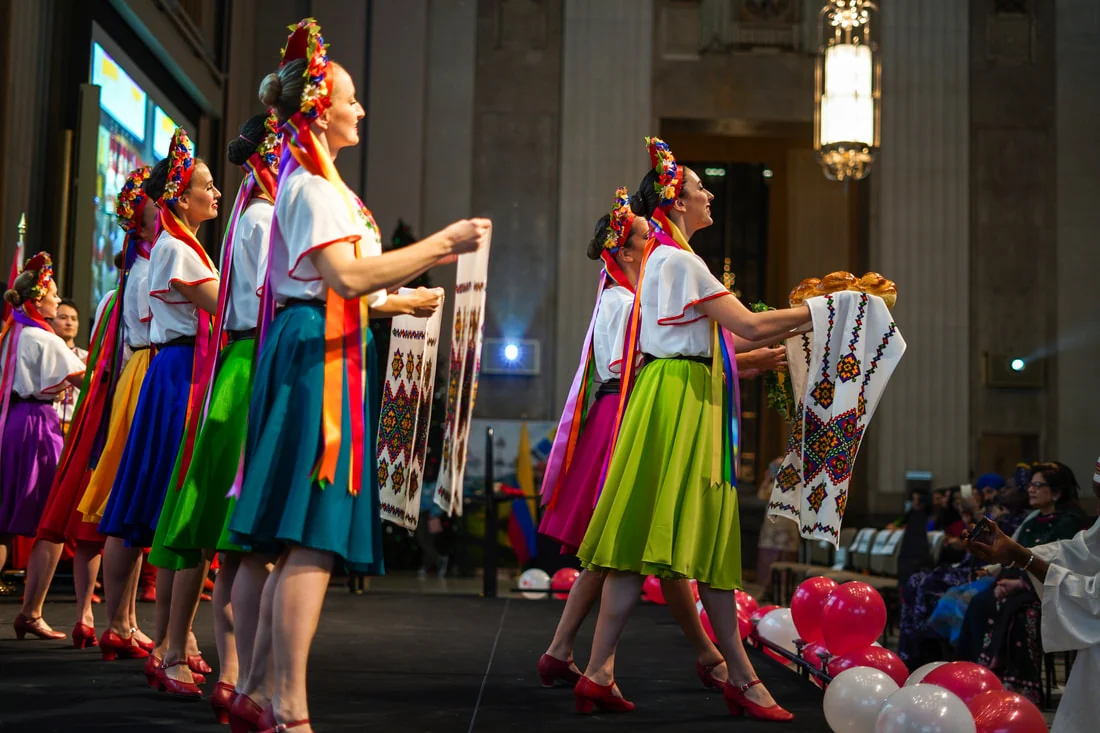 Dancers in bright skirts holding embroidered rushnyky, one carrying bread, in a welcome dance inside a stone hall