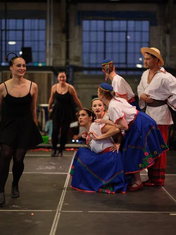 Dancers in blue skirts and embroidered shirts crouched in a group pose beside a dancer in black rehearsal clothes