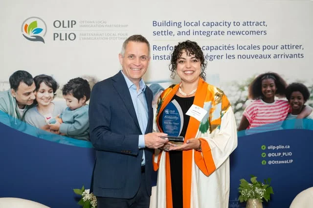 An award being presented in front of an Ottawa Local Immigration Partnership (OLIP/PLIO) backdrop at the launch of Welcoming Ottawa Week 2026