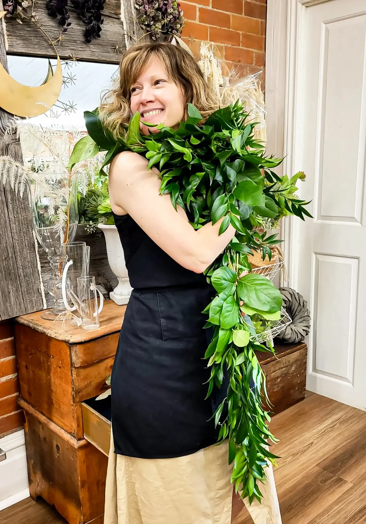 A florist in a black apron laughing as she carries a long garland of glossy greenery over one shoulder inside the shop — the photograph on the shop's About page.