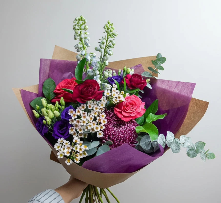 A hand-tied bouquet of red and pink roses, white spirea, purple lisianthus and eucalyptus, wrapped in purple tissue and kraft paper and held up by the stems.