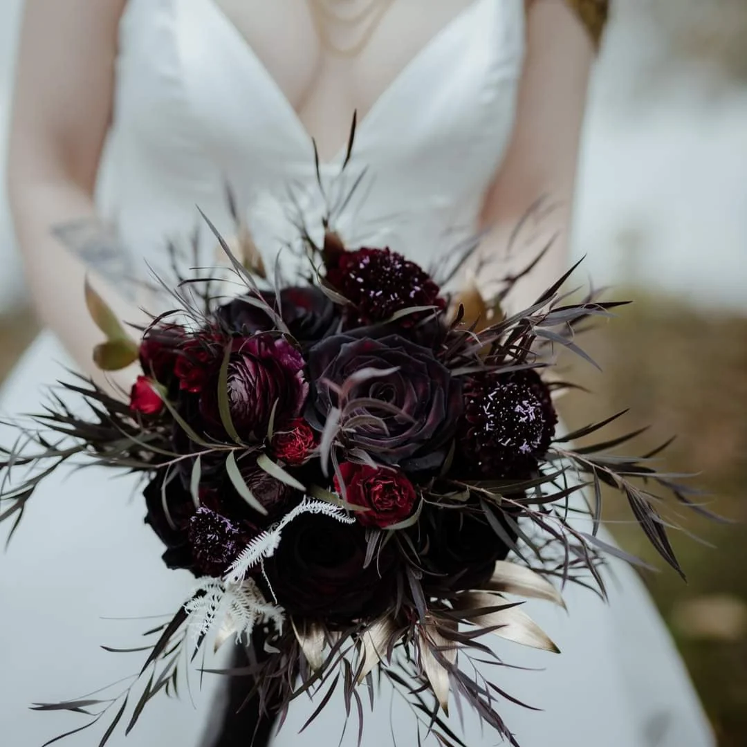 A dark bridal bouquet — burgundy ranunculus, near-black roses and spiky dyed foliage — held in front of a white gown, from the shop's wedding page.