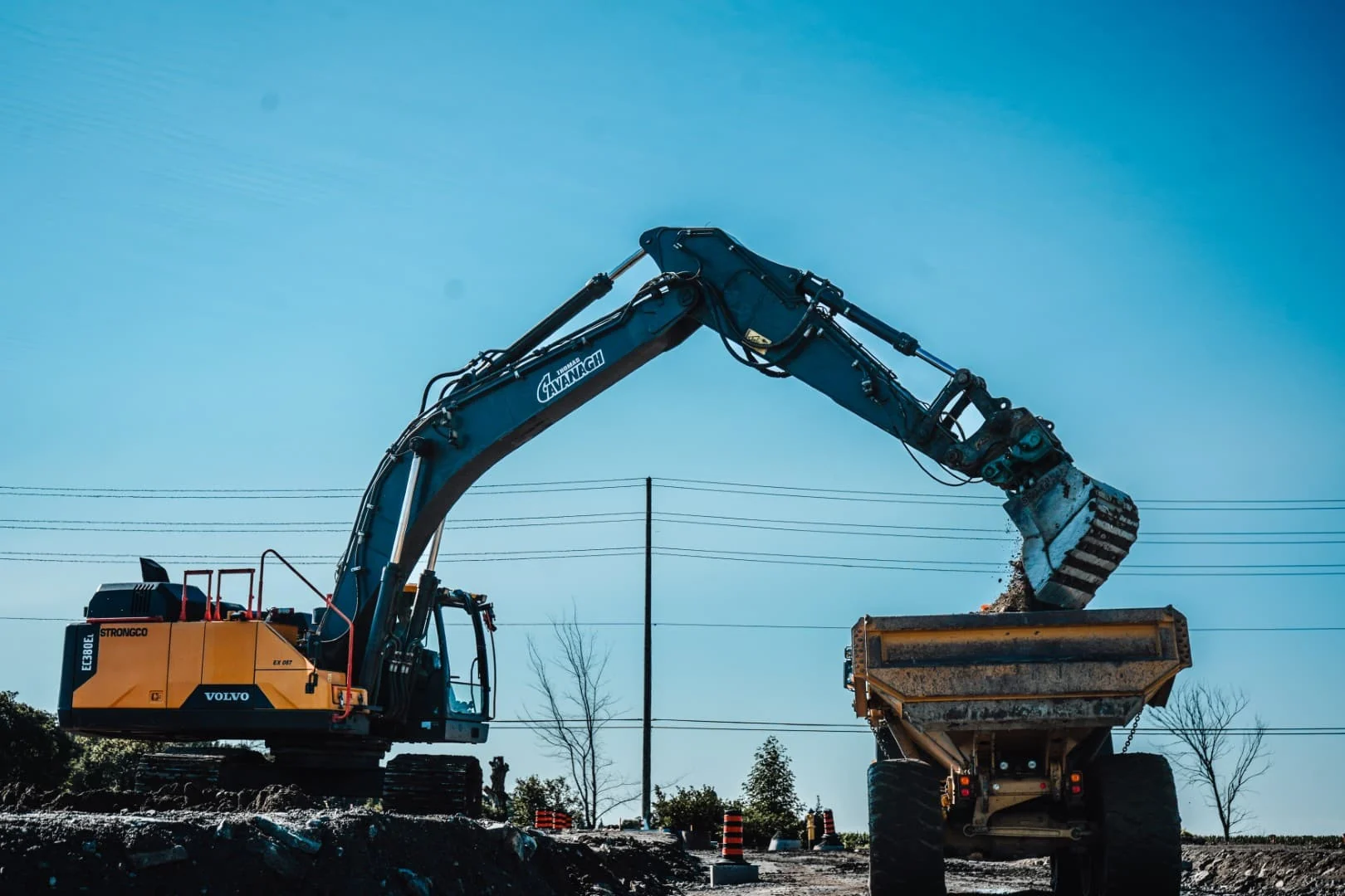 A Volvo tracked excavator with the Cavanagh name on its boom tipping a bucket of earth into an articulated haul truck