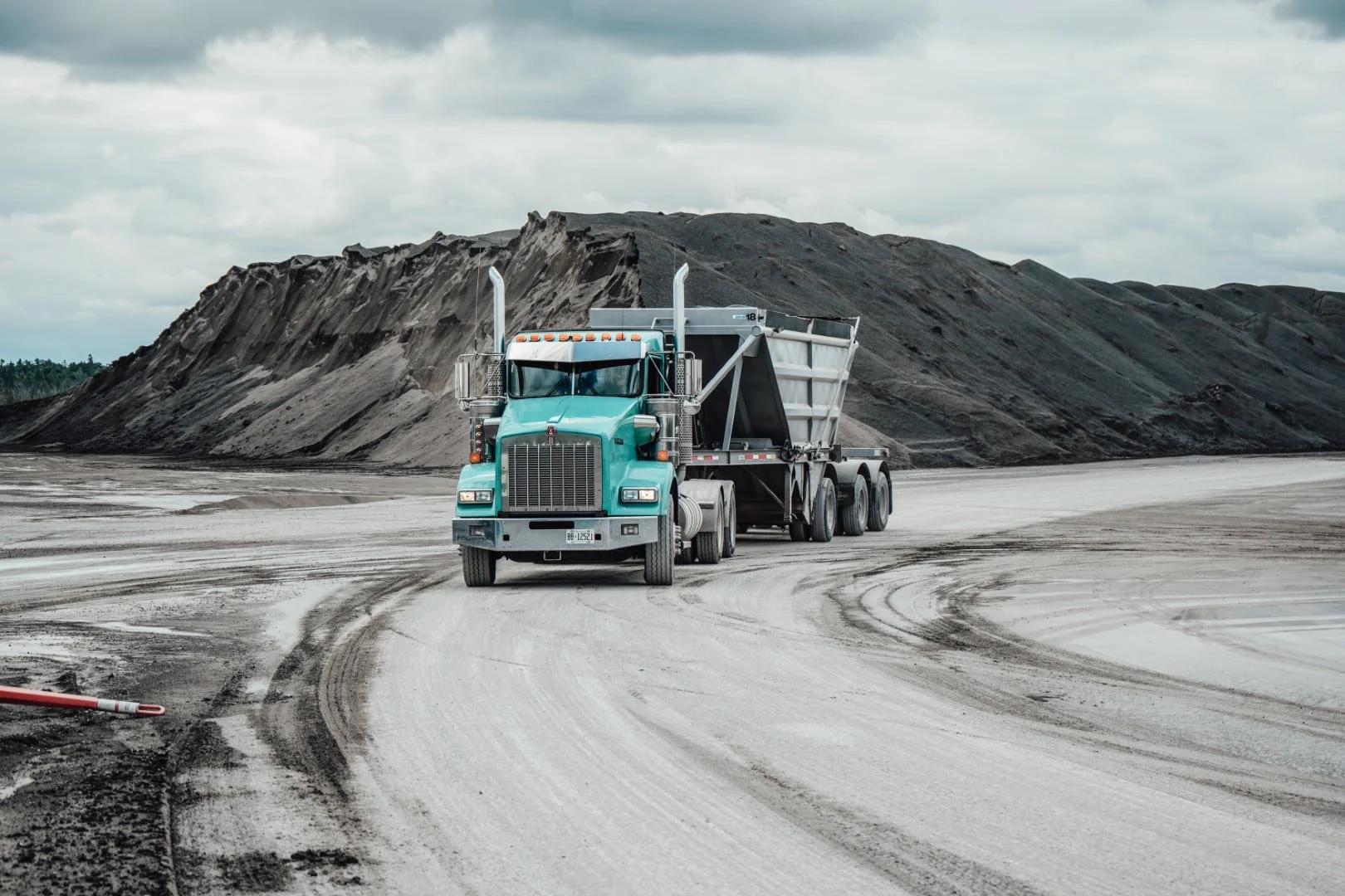 A turquoise Kenworth tractor hauling a dump trailer across a quarry floor, a long dark stockpile behind it under an overcast sky