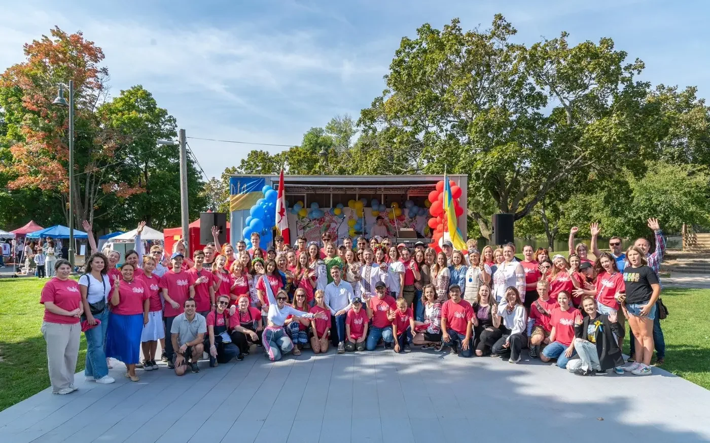 The full UAFest volunteer crew in matching red T-shirts posing and waving together in front of the Main Stage at Riverside Park