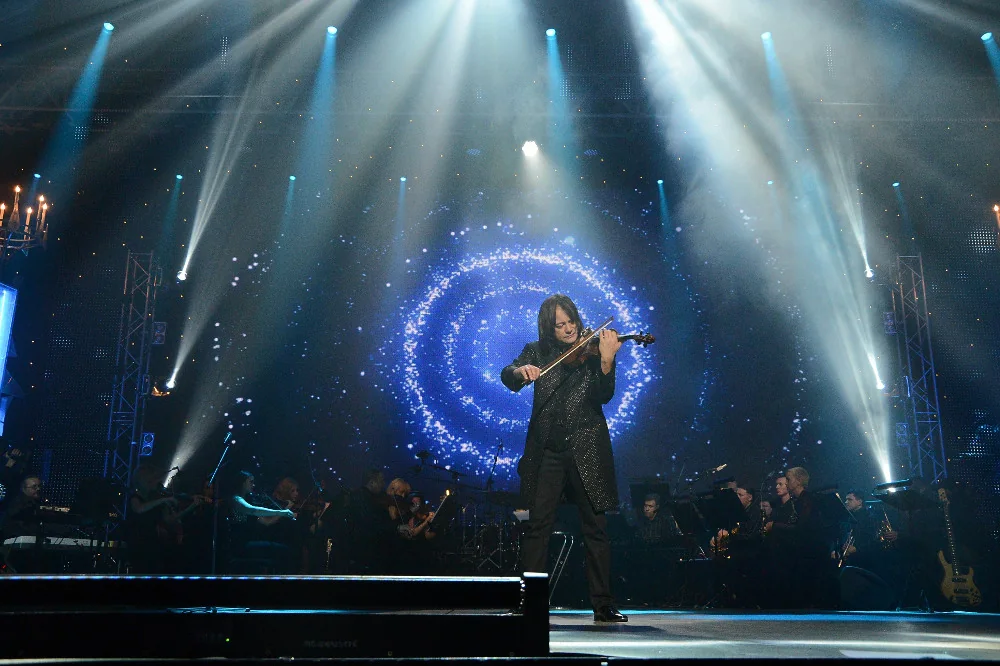 Vasyl Popadiuk alone at the front of a large stage, lit by spotlight beams, orchestra behind him